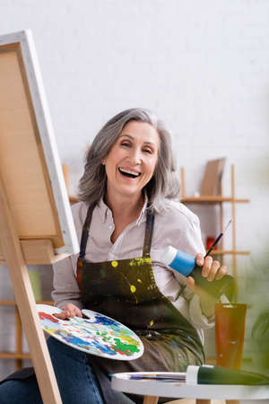 happy mature artist holding tube with blue paint and palette while sitting near canvasの写真素材