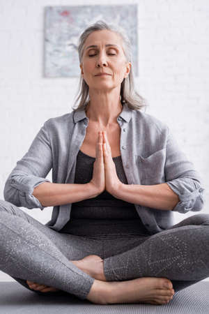mature woman with praying hands and closed eyes sitting in lotus pose on yoga matの写真素材