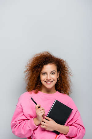 curly woman with notebook and pen smiling at camera isolated on gray