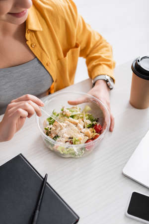 partial view of freelancer holding plastic fork near fresh salad and coffee to goの写真素材