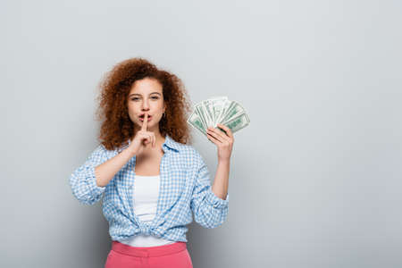 curly woman showing hush gesture while holding dollars on gray background