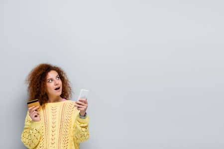 surprised woman looking away while holding smartphone and credit card isolated on gray