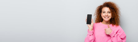 cheerful woman showing thumb up while holding cellphone with blank screen isolated on gray, bannerの写真素材