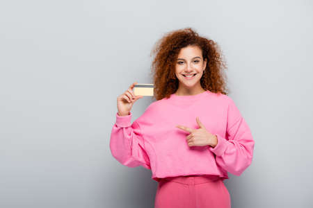 cheerful woman with curly hair pointing at credit card on gray background