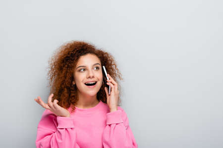 amazed woman looking away while talking on smartphone isolated on grayの写真素材