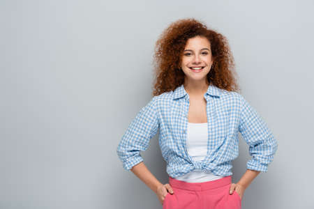 happy woman with wavy hair standing with hands in pockets on gray backgroundの写真素材