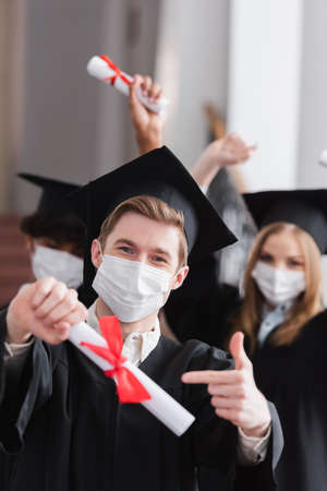 Graduate in medical mask pointing at blurred diplomaの写真素材