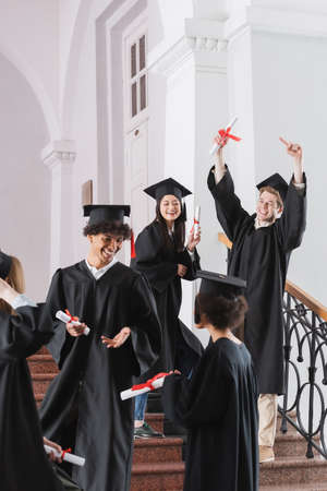 Cheerful student pointing at diploma near multicultural friends on blurred foregroundの写真素材