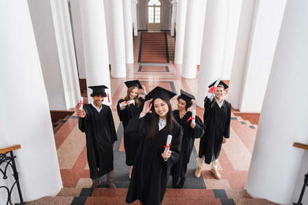 High angle view of cheerful asian graduate holding diploma near friends in capsの写真素材