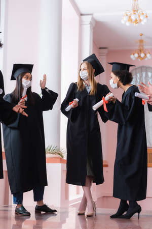 Multiethnic graduates in medical masks holding diplomas and talking in hall of universityの写真素材
