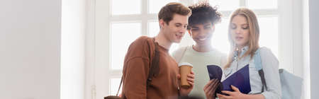 Interracial students with coffee to go and notebook standing near window, bannerの写真素材