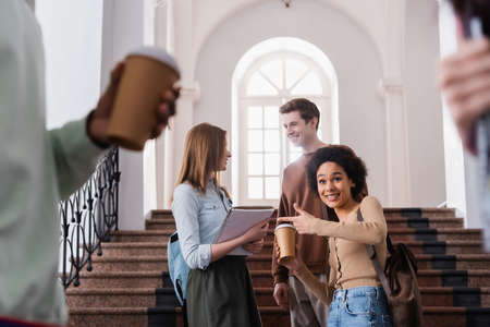 Smiling african american student with coffee to go pointing near friends in universityの写真素材