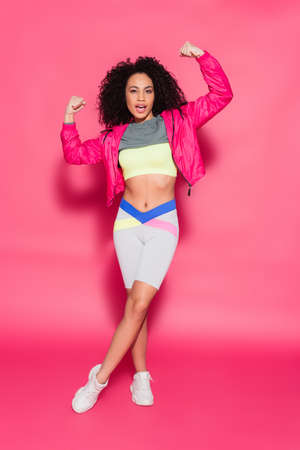 full length of sporty brunette african american woman showing muscles while posing on pink