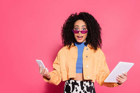 confused african american woman in sunglasses holding smartphone and digital tablet on pink