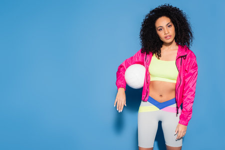 curly african american woman in pink jacket and crop top standing with volleyball on blueの写真素材