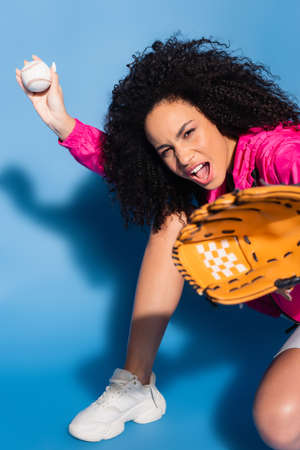 excited african american woman in leather glove holding baseball on blueの写真素材