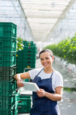 cheerful african american farmer standing with digital tablet near plastic boxes in greenhouse