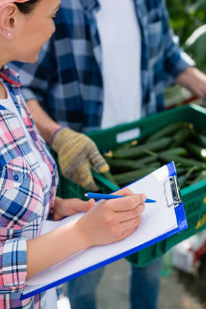 african american farmer writing on clipboard near colleagues with box of fresh cucumbers on blurred...