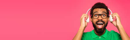 excited african american man in glasses holding wireless headphones isolated on pink, banner