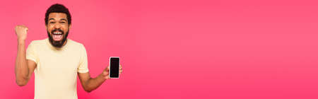 excited african american man holding smartphone with blank screen isolated on pink, bannerの写真素材