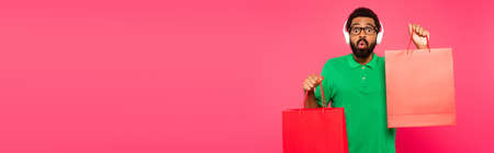 shocked african american man in glasses and headphones holding shopping bags on pink, bannerの写真素材