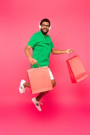 full length of excited african american man in glasses and headphones holding shopping bags on pinkの写真素材