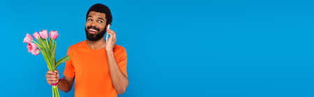 cheerful african american man holding pink tulips and talking on smartphone isolated on blue, bannerの写真素材