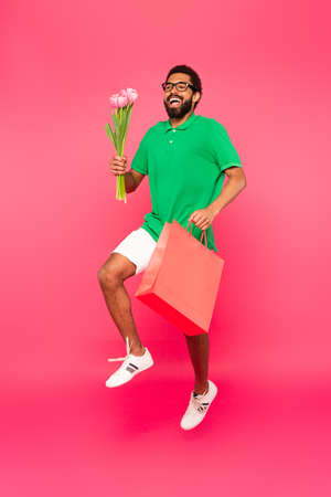 full length of happy african american man in glasses holding shopping bag and bouquet of tulips on pinkの写真素材