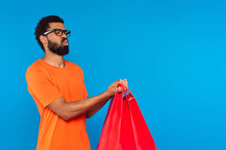 african american man in glasses holding shopping bags and pouting lips isolated on blueの写真素材
