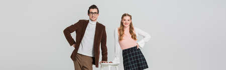 joyful couple in retro clothes posing with hands on hips near high stool isolated on gray, banner