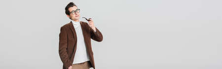 old fashioned man looking at camera while posing with smoking pipe isolated on gray, banner