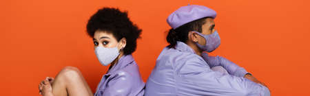 trendy african american couple in protective masks sitting isolated on orange, bannerの写真素材