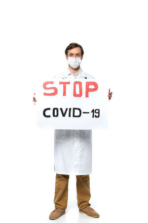 Doctor in white coat and protective mask holding placard with stop covid-2019 lettering on white backgroundの写真素材