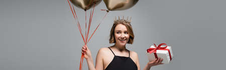 happy plus size woman in slip dress and crown holding golden balloons and present isolated on gray,...