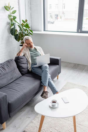 man with gray hair sitting on couch and using laptop in living room