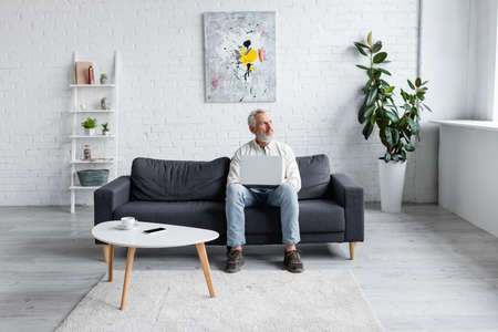 bearded man with laptop sitting on couch near cup of coffee and smartphone with blank screen on coffee tableの写真素材