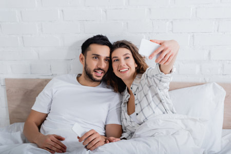 happy woman taking selfie with bearded muslim man in bedの写真素材