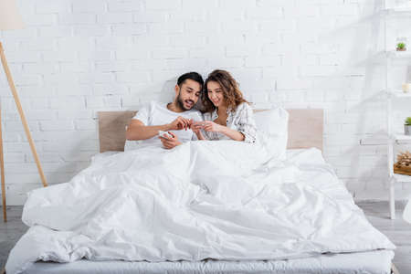 interracial couple smiling while lying on bed and using smartphonesの写真素材