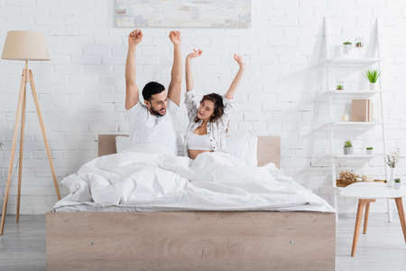 awake interracial couple looking at each other while stretching in bedの写真素材