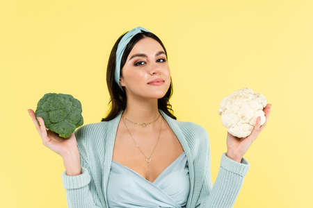 young woman smiling at camera while holding cauliflower and broccoli isolated on yellowの写真素材