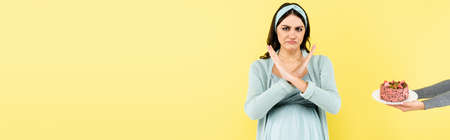 displeased pregnant woman showing reject gesture near cake isolated on yellow, bannerの写真素材
