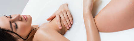 young woman relaxing in bath with milk while covering breast with hand, bannerの写真素材