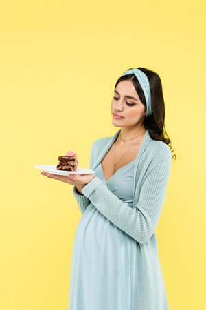 young pregnant woman feeling doubt while holding delicious cake isolated on yellowの写真素材