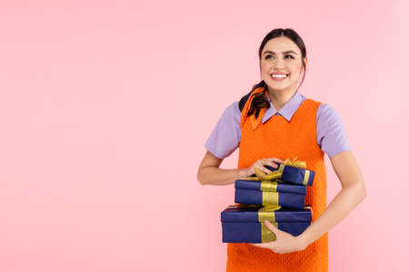 excited woman looking away while holding gift boxes isolated on pinkの写真素材