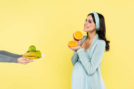 joyful pregnant woman holding half of juicy orange near fresh fruits isolated on yellow