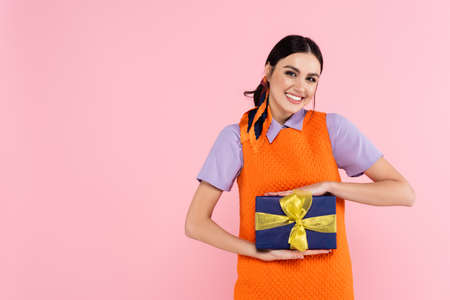 cheerful woman showing present while looking at camera isolated on pink