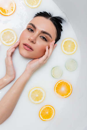 young woman touching face and looking at camera in milk bath with citrus slicesの写真素材