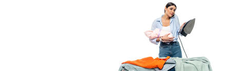 discouraged woman talking on mobile phone while holding infant and iron isolated on white, bannerの写真素材