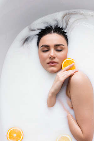 young woman with closed eyes holding orange slice while relaxing in milk bathの写真素材