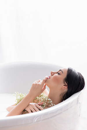 young woman touching chin and holding gypsophila flowers while bathing in milk